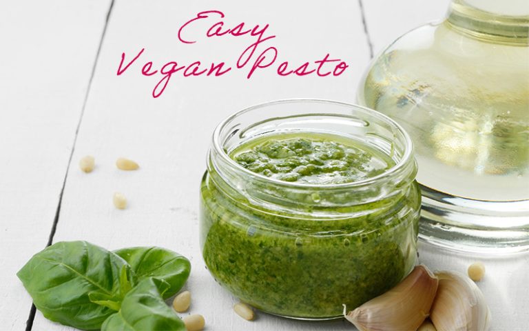 Basil pesto in a glass jar on white surface with basil leaves, pine nuts, garlic and olive oil in background